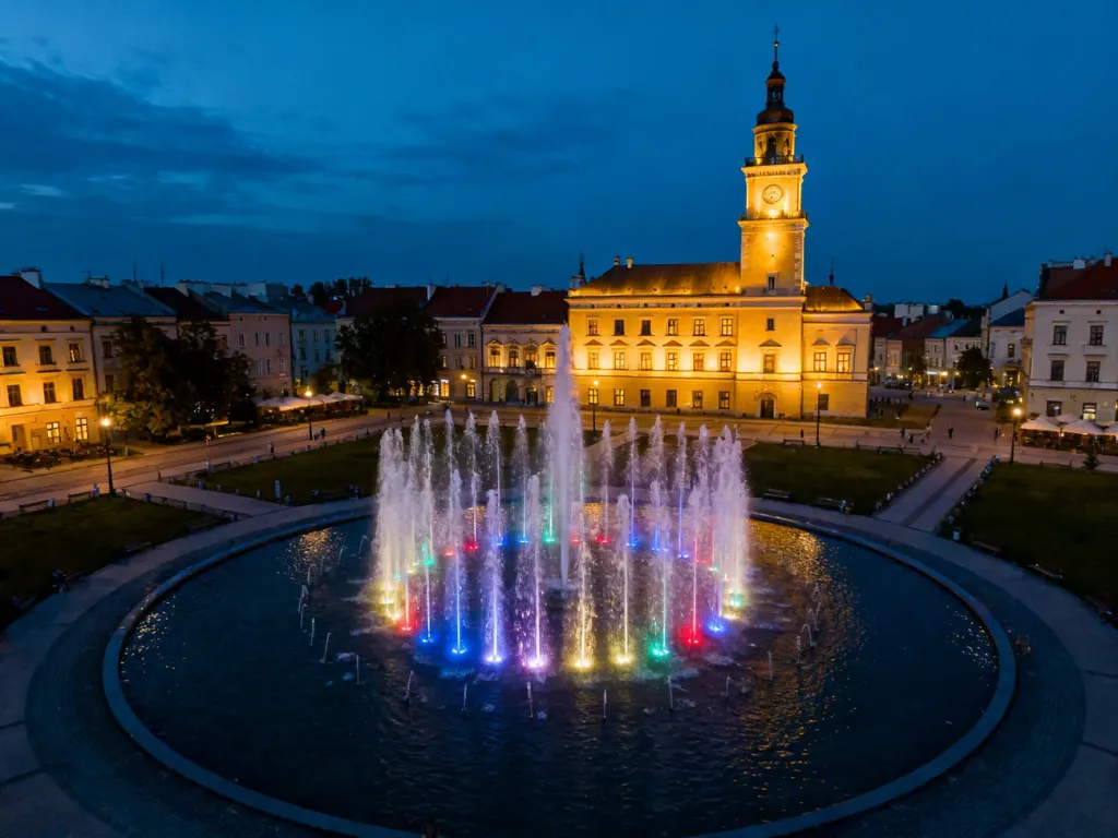 Wieczorna iluminacja fontanny multimedialnej w Rzeszowie z widocznym w tle zabytkowym ratuszem.
